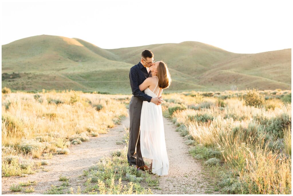 couple kissing on dirt road in idaho with rolling foot hills in the sunset. photo taken by Miranda Renee Photography
