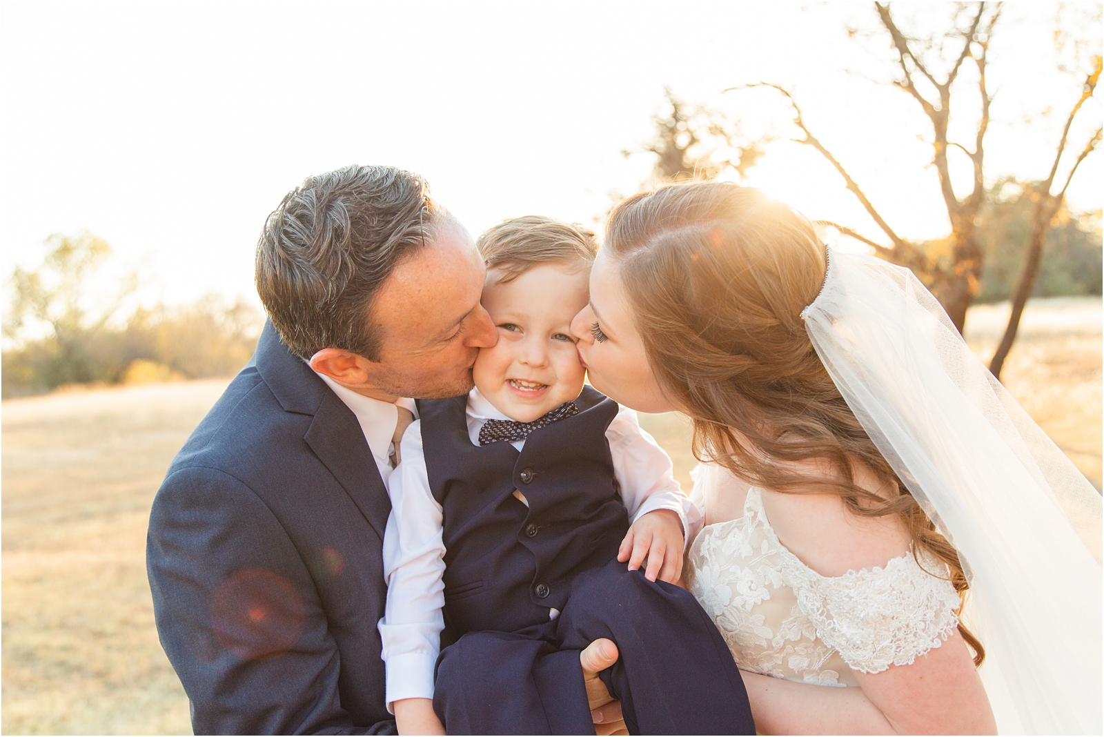bride and groom kissing their son on the cheeks with a sunset it the background
